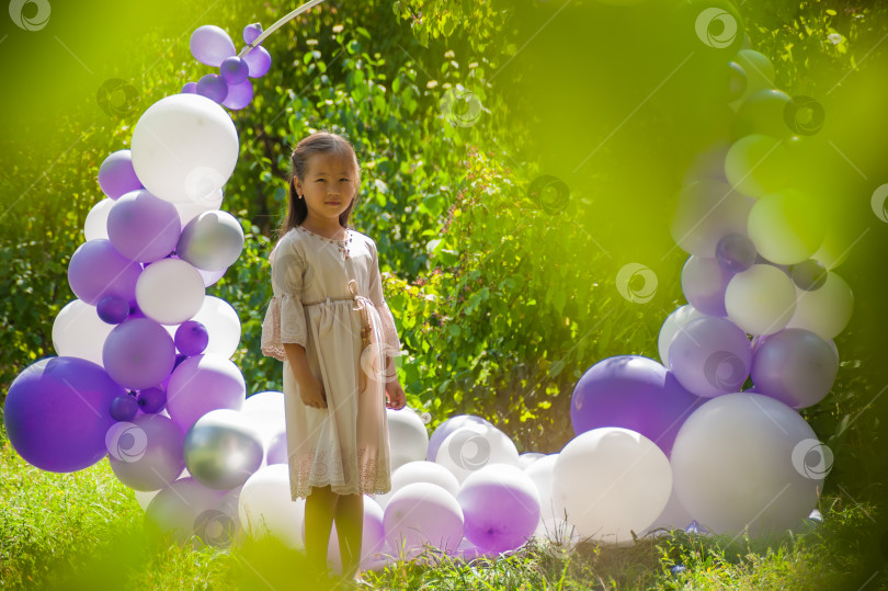 Скачать 7-летняя азиатская девочка позирует в платье среди зелени на фоне арки из воздушных шаров фотосток Ozero