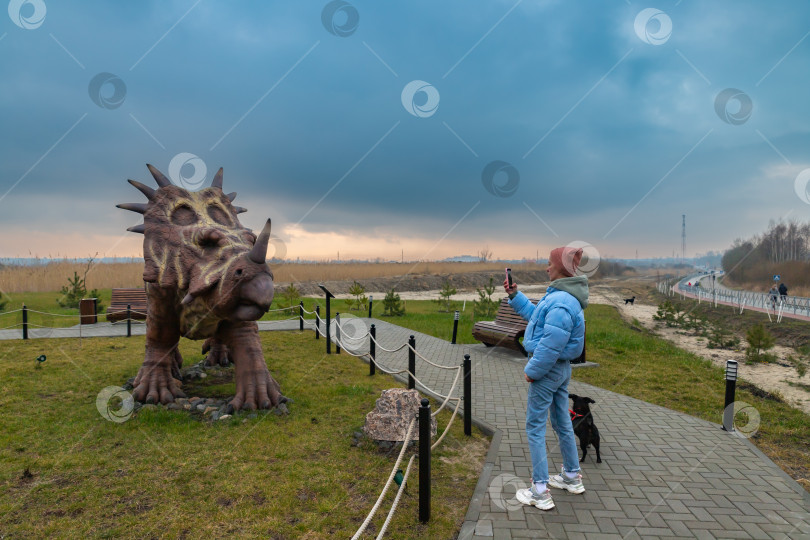 Скачать Девушка с собакой фотографирует фигуру стиракозавра в Динопарке Янтарного фотосток Ozero