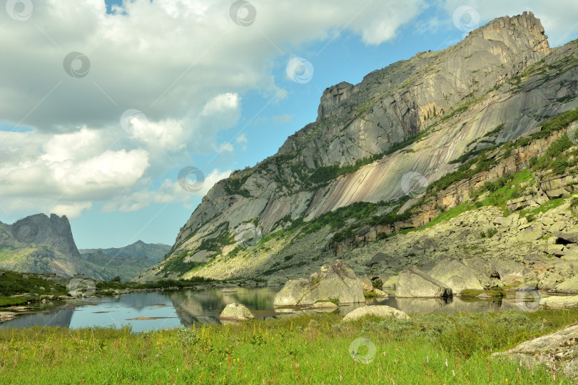 Скачать Высокий и отвесный скальный массив нависает над широким, спокойным горным озером в пасмурный летний день. фотосток Ozero