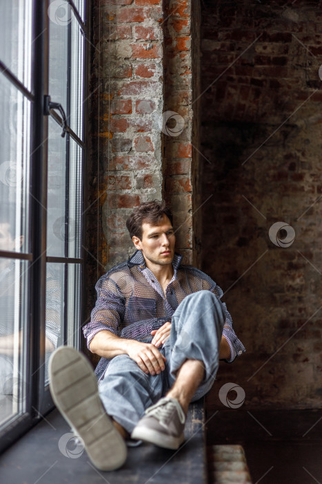 Скачать Современный молодой человек со стильной прической сидит на подоконнике у окна на темном фоне фотосток Ozero