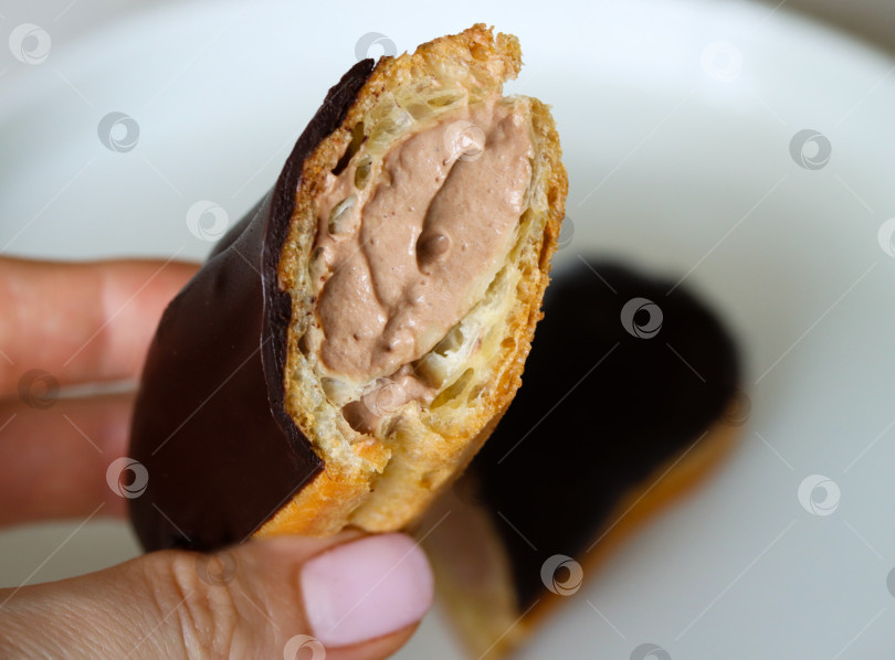 Скачать Рука, держащая шоколадный эклер, демонстрирует его кремовую начинку. Крупный план наполовину съеденного шоколадного эклера в руке женщины. Эклер в шоколаде с обильной начинкой, крупный план фотосток Ozero