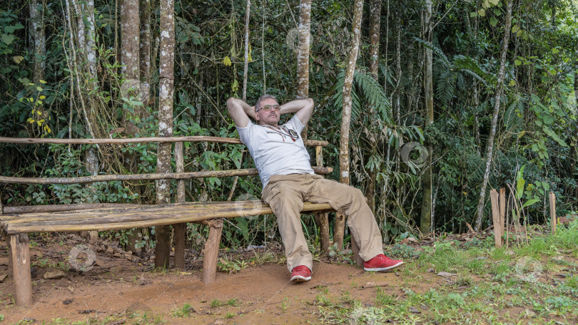 Скачать Мужчина отдыхает, сидя на некрашеной деревянной скамье на поляне в тропическом лесу. фотосток Ozero