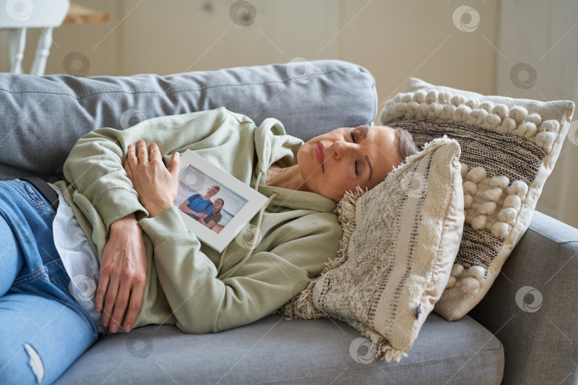 Скачать Грустная зрелая женщина держит в руках семейную фотографию, лежа с закрытыми глазами на диване у себя дома фотосток Ozero