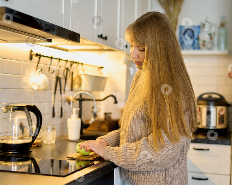 Скачать Белокурая женщина режет зеленое яблоко на доске на кухне. Полезная закуска в виде ломтиков фотосток Ozero