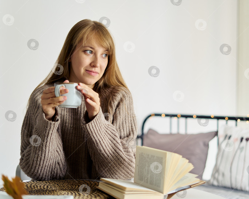 Скачать Счастливая молодая девушка в свитере мечтает с чашкой чая, читая книгу в спальне фотосток Ozero