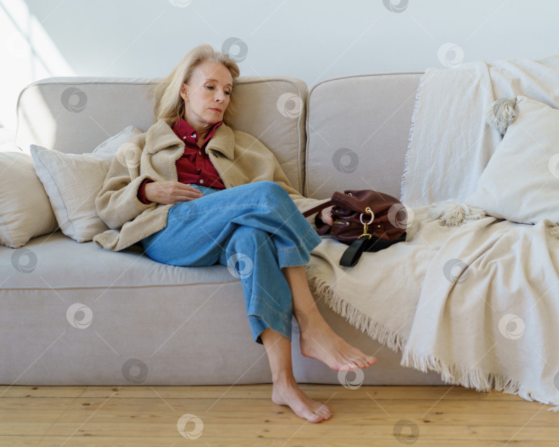 Скачать Усталая пожилая женщина смотрит в сторону, отдыхая дома на диване после тяжелого рабочего дня фотосток Ozero