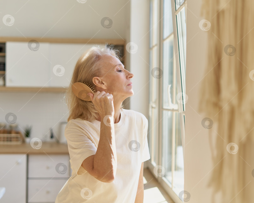 Скачать Красивая пожилая женщина расчесывает волосы и смотрит в окно, проводя свободное время дома фотосток Ozero