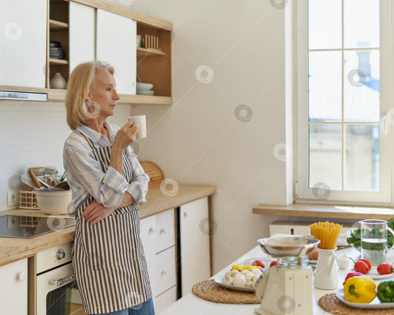 Скачать Задумчивая пожилая женщина, начинающая новый день с кофе, стоя дома на кухне фотосток Ozero