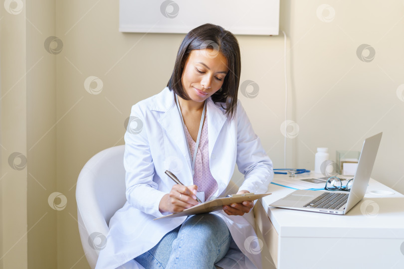 Скачать Молодая африканка-врач в белом халате сидит на рабочем месте в клинике и заполняет анкету пациента фотосток Ozero