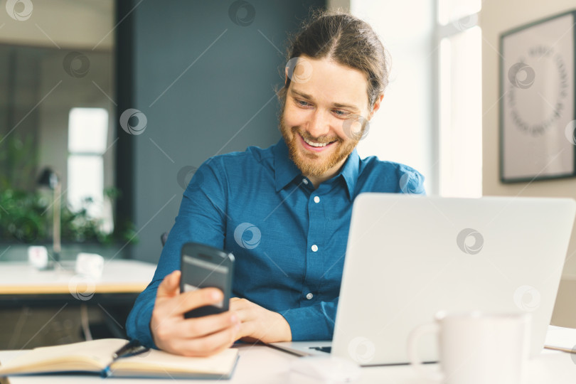 Скачать Улыбающийся молодой человек в повседневной одежде смотрит на экран смартфона на рабочем месте фотосток Ozero