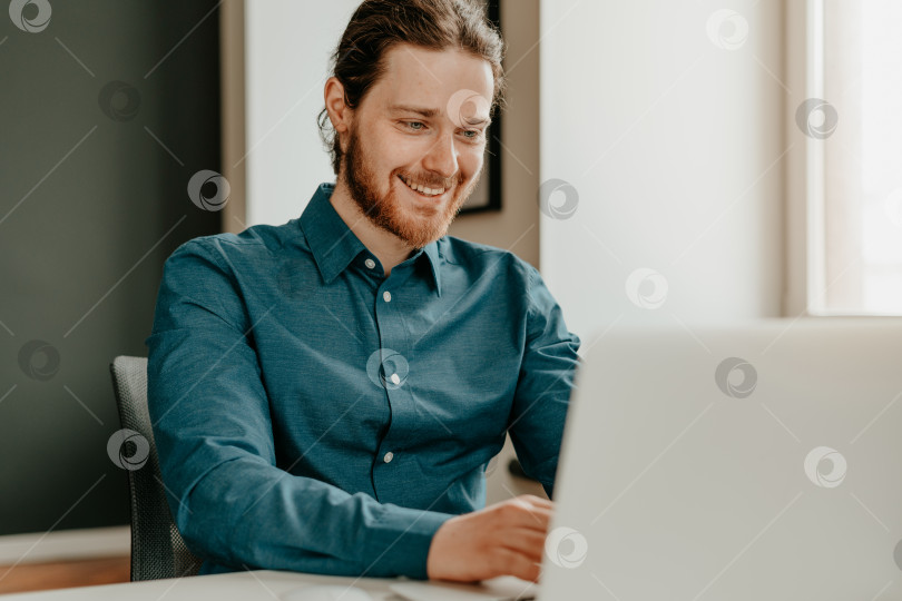 Скачать Улыбающийся молодой офисный работник мужского пола печатает на клавиатуре и смотрит на экран ноутбука в офисе. фотосток Ozero