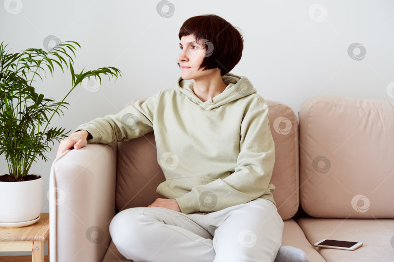Скачать Зрелая женщина, проводящая время дома, сидит в комнате на диване и выглядит грустной. Концепция цифровой детоксикации фотосток Ozero
