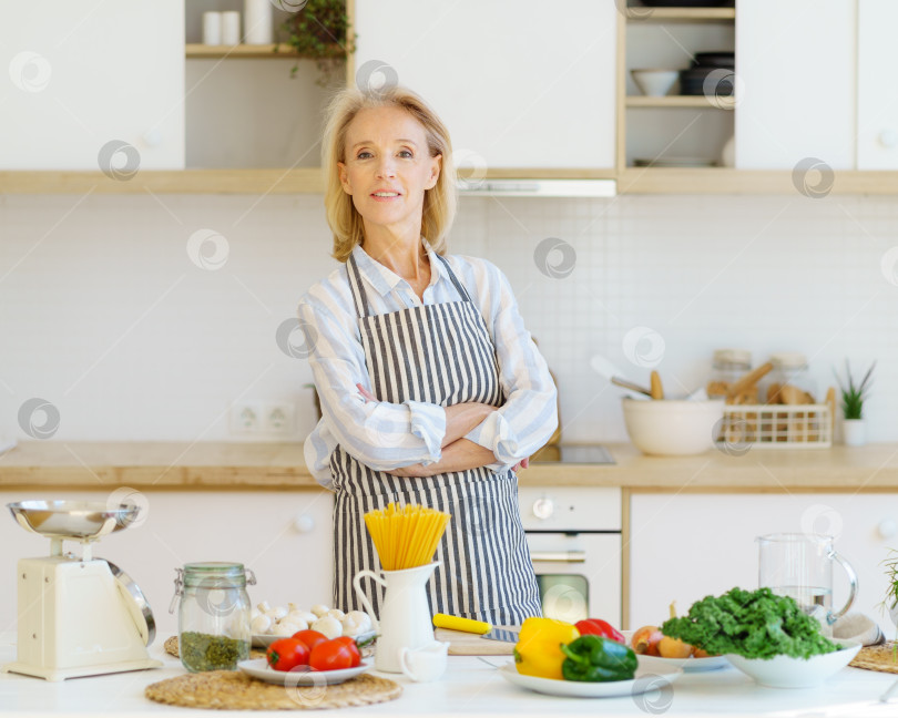 Скачать Портрет красивой пожилой женщины, стоящей на кухне и улыбающейся в камеру во время приготовления здоровой пищи фотосток Ozero