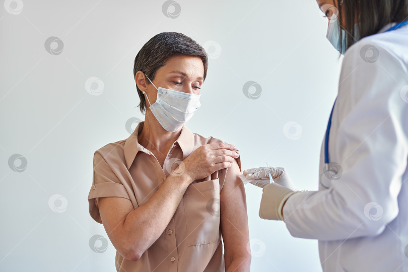 Скачать Женщина средних лет в маске для лица сидит в кабинете врача и делает прививку от covid-19 во время пандемии фотосток Ozero