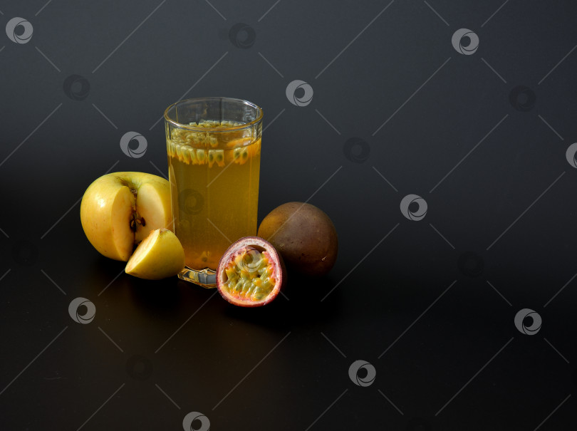 Скачать Высокий стакан фруктового сока с семенами на черном фоне, рядом с кусочками спелой маракуйи и зеленым яблоком. фотосток Ozero