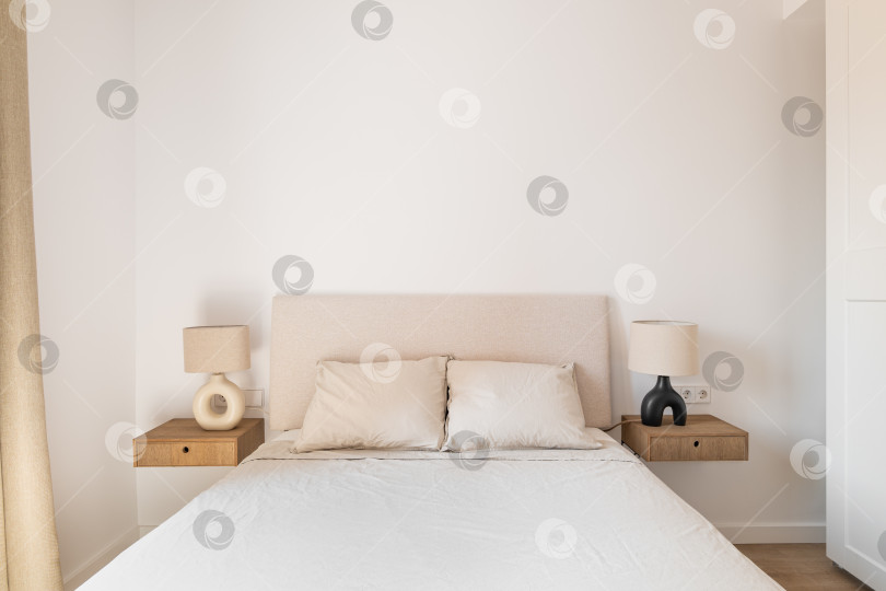 Скачать Горизонтальный снимок двуспальной кровати с белыми подушками и покрывалом, прикроватными тумбочками, бра на фоне белых обоев и шкафа-купе. Концепция новой квартиры для молодой семьи фотосток Ozero