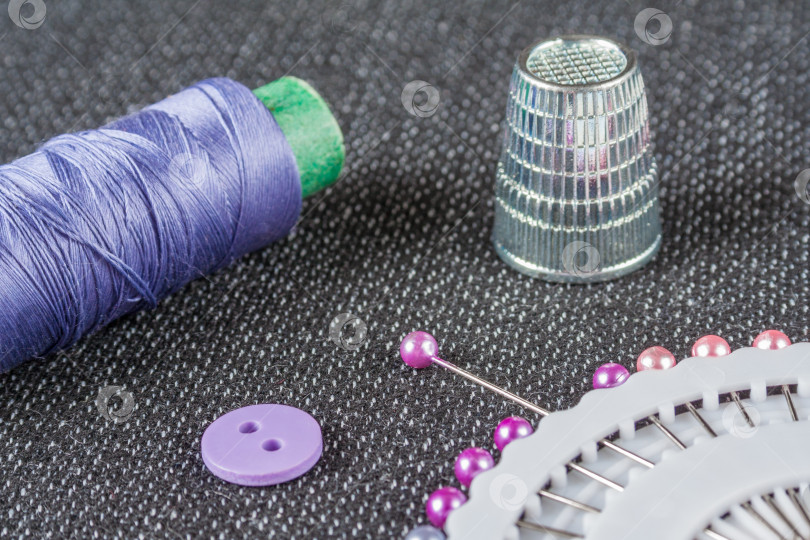 Скачать Швейные принадлежности: фиолетовая катушка с нитками, иголки, наперсток, пуговица на фактурной поверхности ткани фотосток Ozero