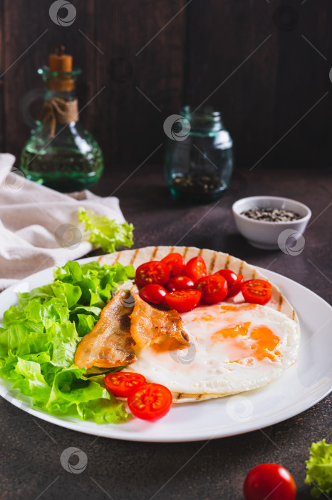 Скачать Аппетитные тако с жареным яйцом, беконом, помидорами и листьями салата на тарелке на столе, вид сверху фотосток Ozero