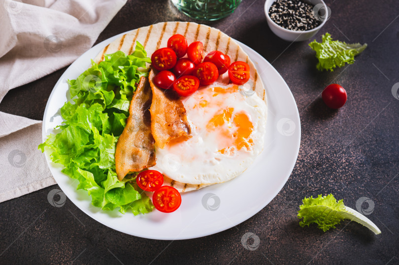 Скачать Тако на гриле с яйцом, жареным беконом, овощами и зеленью на тарелке на столе фотосток Ozero