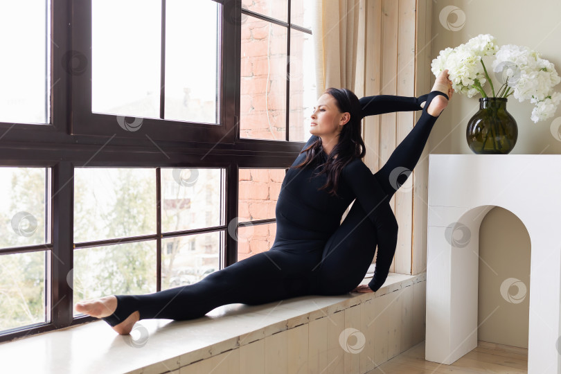 Скачать Брюнетка выполняет упражнения йоги дома: Паривритта Сурья Янтрасана, поза циркуля, тренировка в спортивной одежде, сидя на подоконнике фотосток Ozero