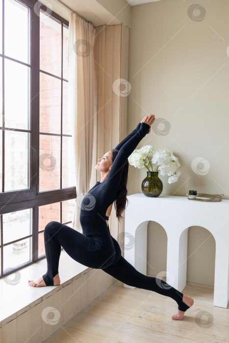 Скачать Женщина, практикующая йогу, выполняет упражнение Вирабхадрасана, поза воина, стоит у окна и опирается одной ногой на подоконник, тренируется в черном спортивном костюме, вид в полный рост фотосток Ozero