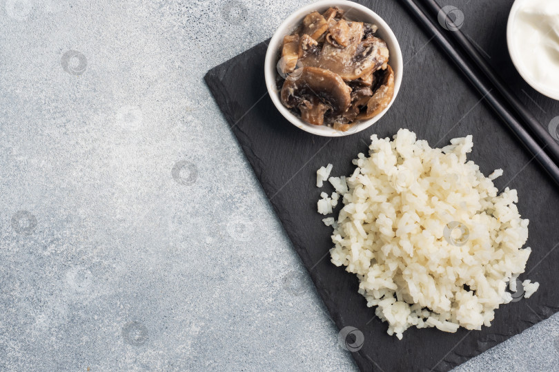 Скачать Отварной белый рис и грибы в сметане на черной подставке с палочками для еды. Серый бетонный фон копирует пространство. фотосток Ozero