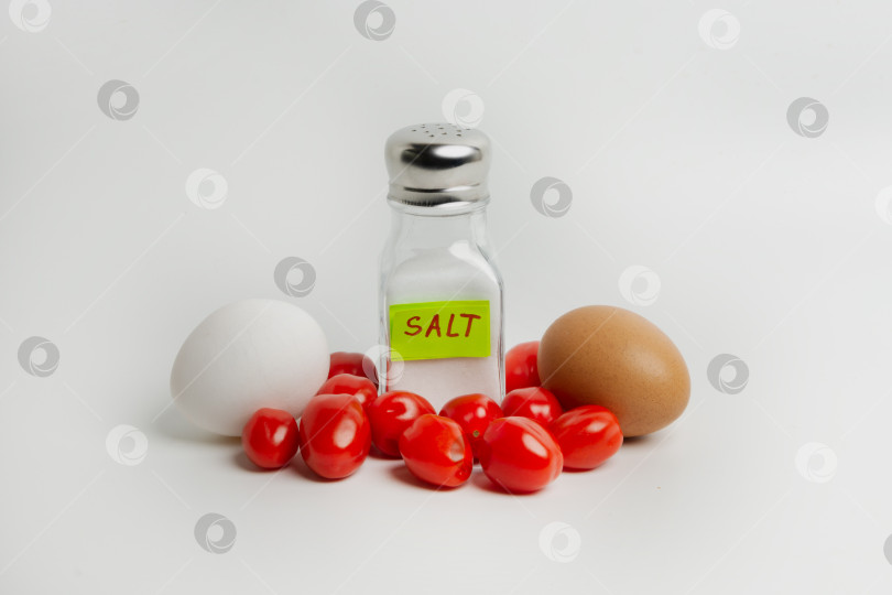 Скачать Яйца, помидоры и соль в солонке на белом фоне. Пересаливать продукты вредно для здоровья. Солить пищу нужно в меру фотосток Ozero
