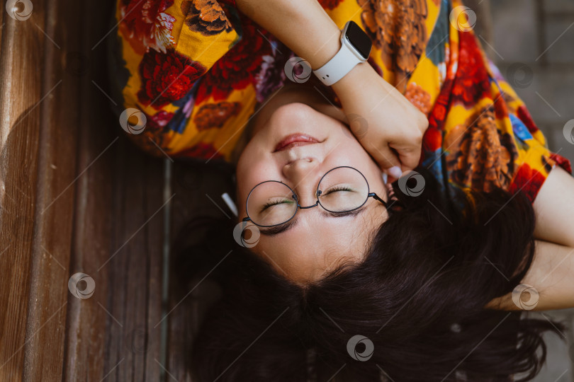 Скачать Расслабленная азиатка слушает музыку в наушниках, лежа на скамейке в парке фотосток Ozero