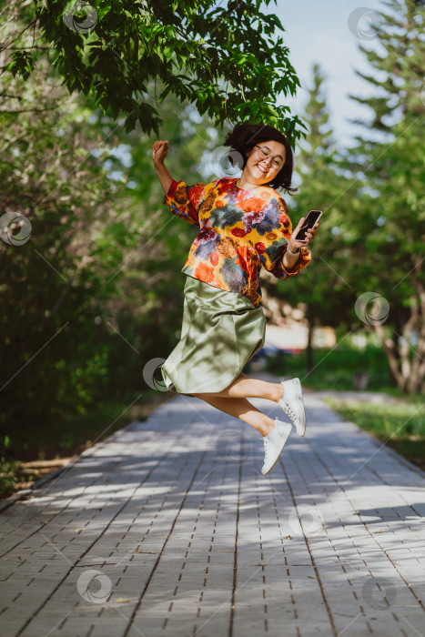 Скачать Взрослая азиатка радостно прыгает со смартфоном в залитом солнцем парке фотосток Ozero