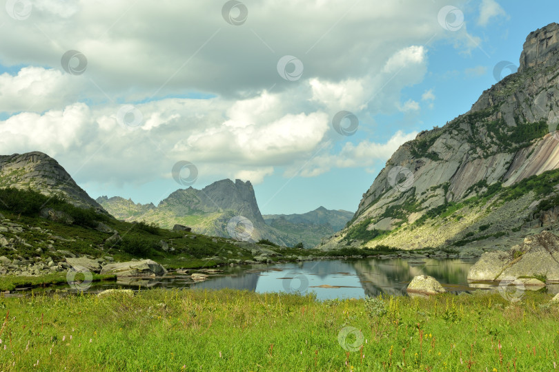 Скачать Небольшая поляна перед спокойным озером с отражением высоких скал, окруженных горными хребтами, в солнечный летний день. фотосток Ozero