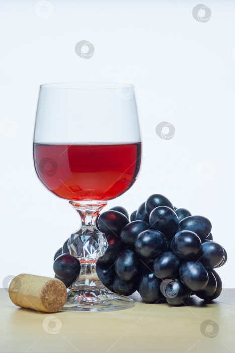 Скачать Бокал красного вина рядом с гроздью черного винограда и пробкой на деревянном столе на белом фоне (концепция) фотосток Ozero