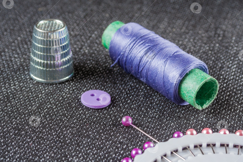 Скачать Швейные принадлежности: фиолетовая катушка с нитками, иголки, наперсток, пуговица на темной фактурной поверхности ткани фотосток Ozero