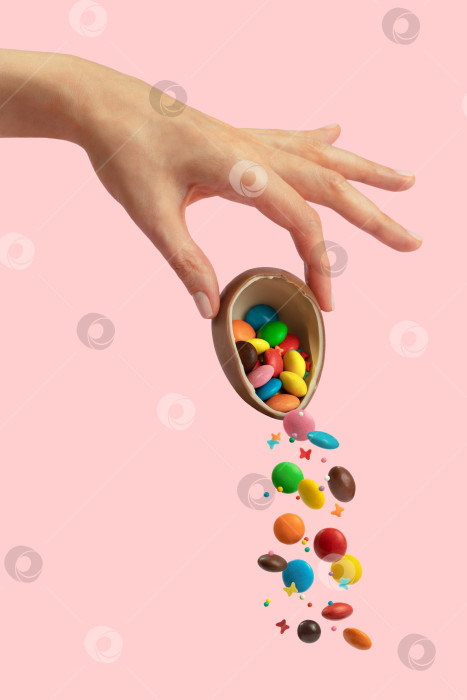 Скачать Женская рука держит шоколадное пасхальное яйцо, из которого высыпаются разноцветные сладости. Пастельно-розовый фон фотосток Ozero