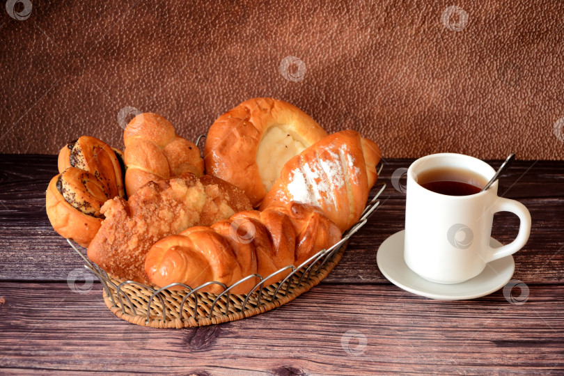 Скачать Чашка горячего чая на блюдце и плетеная корзинка с разнообразной домашней свежей выпечкой на деревянном столе. фотосток Ozero