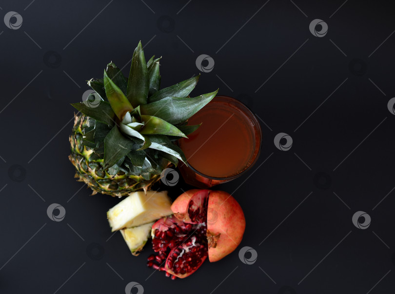 Скачать Высокий стакан фруктового сока на черном фоне, рядом с разбитым гранатом и кусочками спелого ананаса. фотосток Ozero