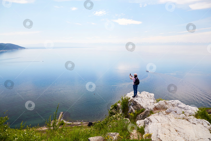 Скачать Путешественница стоит на скале и фотографирует озеро Байкал, деревню Листвянка летним днем фотосток Ozero
