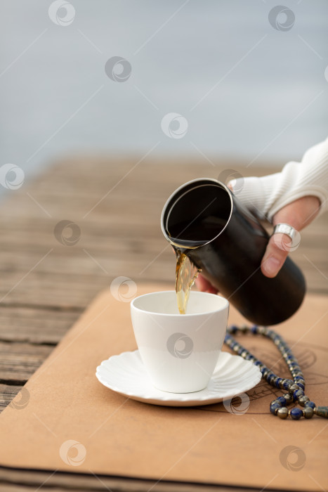 Скачать Женщина после занятий йогой наливает травяной чай из термоса в белую чашку, рядом с ней лежат бусины Малас фотосток Ozero