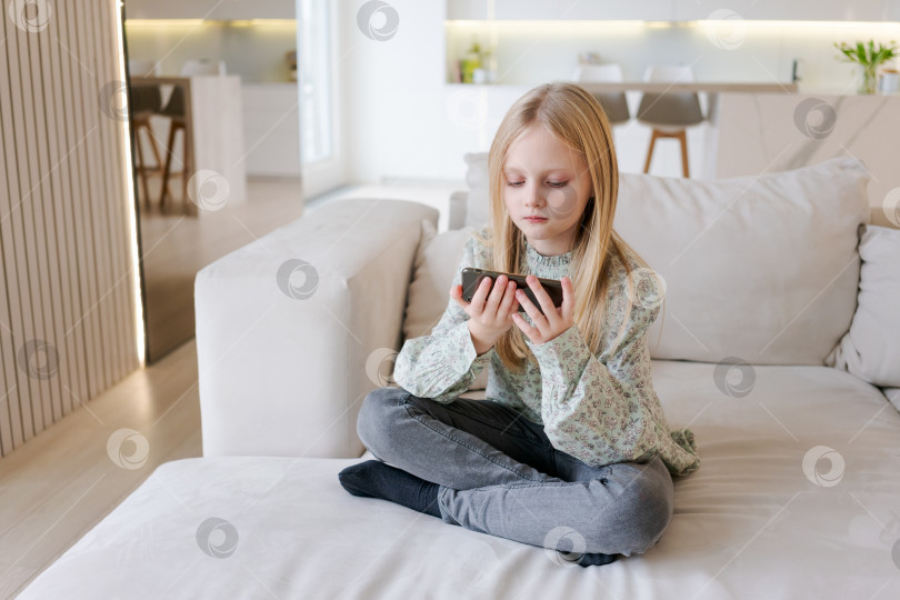 Скачать Молодая девушка сидит на диване и смотрит на свой мобильный телефон фотосток Ozero