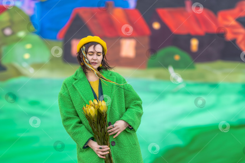 Скачать Молодая женщина в зеленом брючном костюме, пальто и желтом берете стоит с букетом желтых весенних цветов на весеннем фоне. Концепция весеннего праздника - 8 марта, Пасха фотосток Ozero