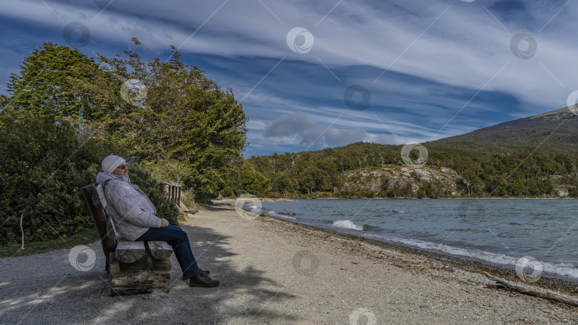 Скачать Мужчина в пуховике сидит, отдыхая, на бревенчатой скамейке у изумрудного озера. фотосток Ozero
