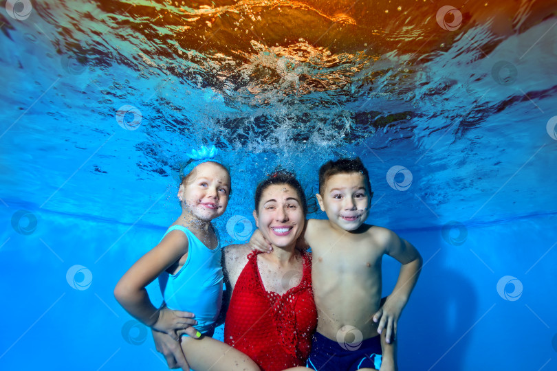 Скачать Красивая, веселая семья: мама, маленькие сын и дочь, плавают и позируют под водой в бассейне в красном и голубом купальниках. Они смотрят в камеру и улыбаются. Концепция семейного отдыха. Портрет. фотосток Ozero