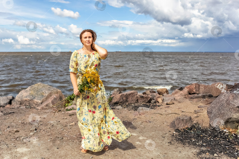 Скачать Молодая женщина в желтом платье с букетом пижмы стоит на пляже на ветру. Легкий и воздушный фотосток Ozero