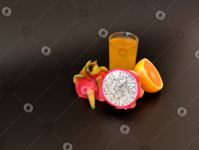 Скачать Стакан желтого сока из экзотических фруктов на черном фоне, рядом с кусочками спелой питайи и апельсина. фотосток Ozero