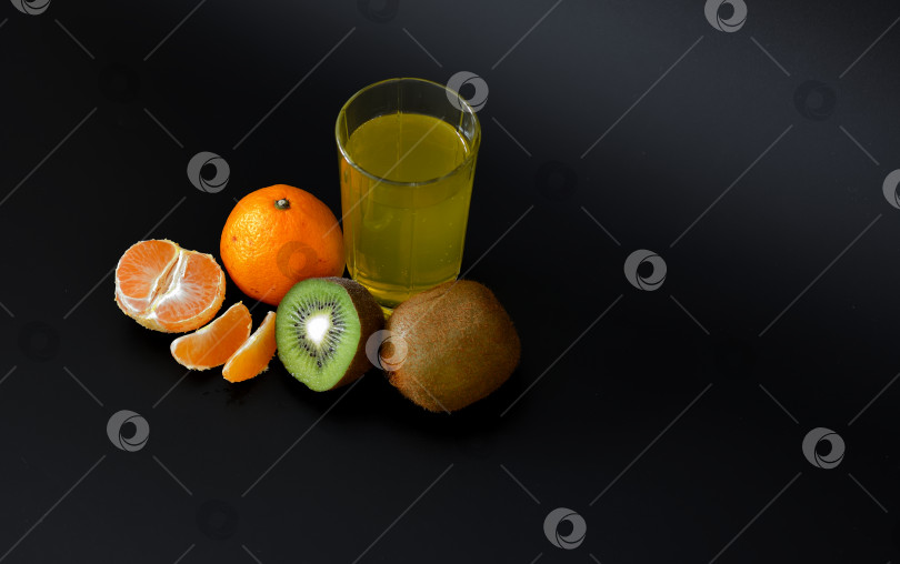 Скачать Стакан сока из тропических фруктов на черном фоне, рядом с кусочками спелых киви и мандаринов. фотосток Ozero