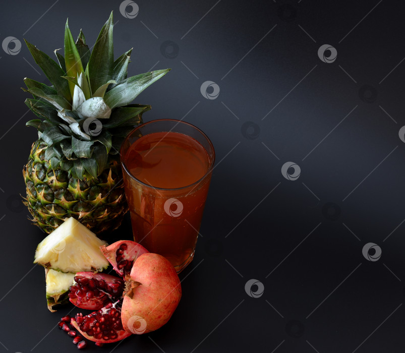 Скачать Стакан свежевыжатого сока на черном фоне, рядом с кусочками спелого ананаса и разломанным плодом граната. фотосток Ozero