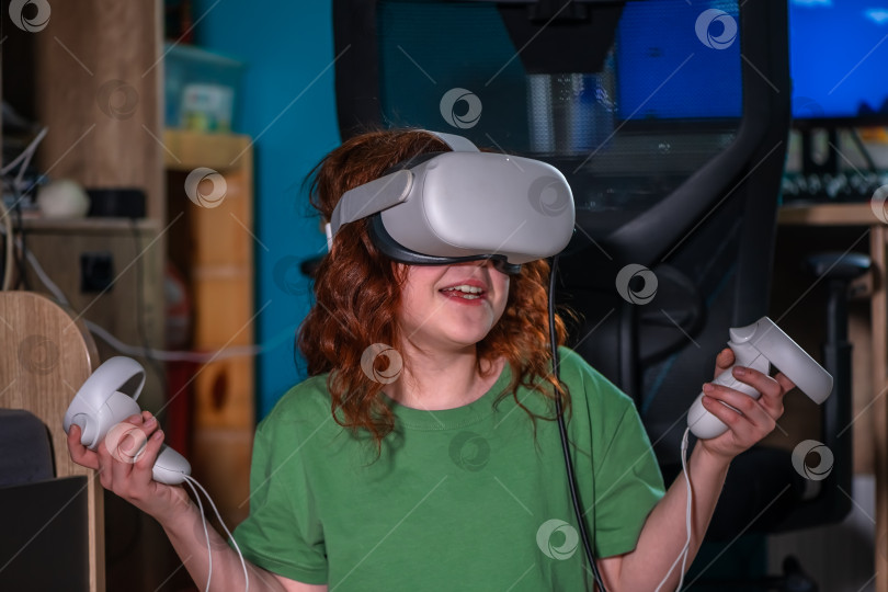 Скачать рыжеволосая девочка-подросток у себя дома в гостиной надела шлем виртуальной реальности, чтобы насладиться онлайн-игрой в киберпространстве. симпатичная девочка развлекается, используя новое электронное устройство для развлечения фотосток Ozero