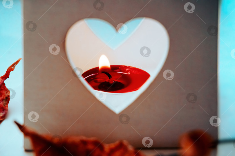 Скачать Квадратный белый подсвечник с вырезанным сердцем и словом love стоит на столе, окруженный сухими осенними листьями, внутри подсвечника горит красная свеча. фотосток Ozero
