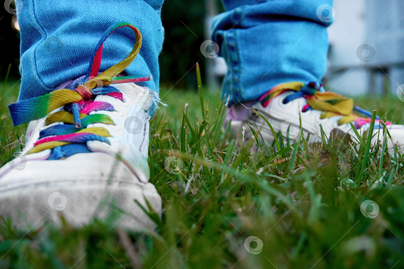 Скачать Ребенок в синих джинсах и белых ботинках с разноцветными шнурками, стоящий на зеленой траве. Селективный селективный мягкий фокус фотосток Ozero