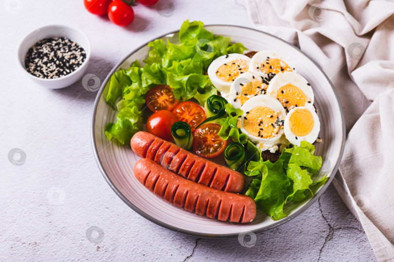 Скачать Тарелка с сосисками, бутербродом с яйцом, помидорами и листьями салата на столе фотосток Ozero