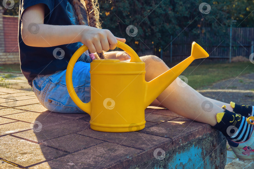 Скачать Девочка сидит на петрушке и придерживает рукой желтую лейку. Инструмент для полива различных видов растений. Избирательный фокус фотосток Ozero